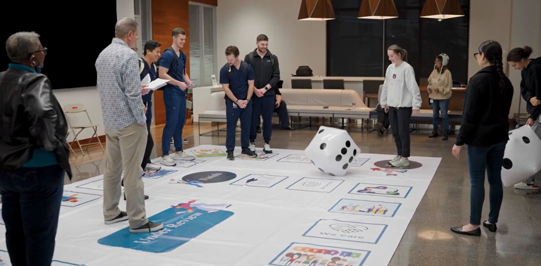 A group of people stands around a large floor board game with a giant dice. 