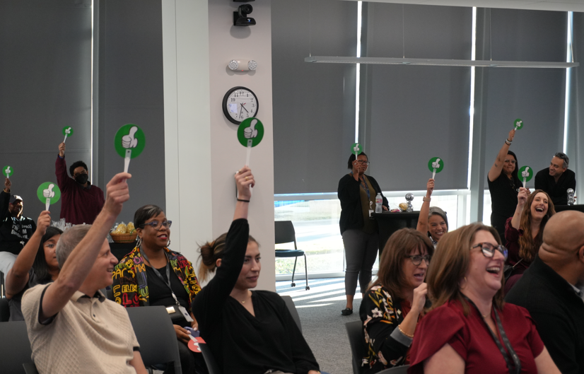 A group of people sitting in a room hold up green thumbs up signs, smiling and laughing. 